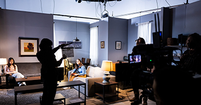 Silhouetted assistant claps the scene board while two female actors wait on couches on a living room set