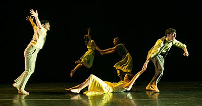Five performers are well lit on a dark stage in dramatic poses and movements