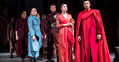 Student actors surround African American male in a bright red suit and robes seeming like Roman times