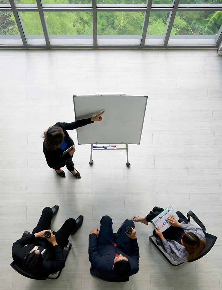 Aerial view of spacious conference room with women in suit presenting to three colleagues by pointing at an easelPlaceholder