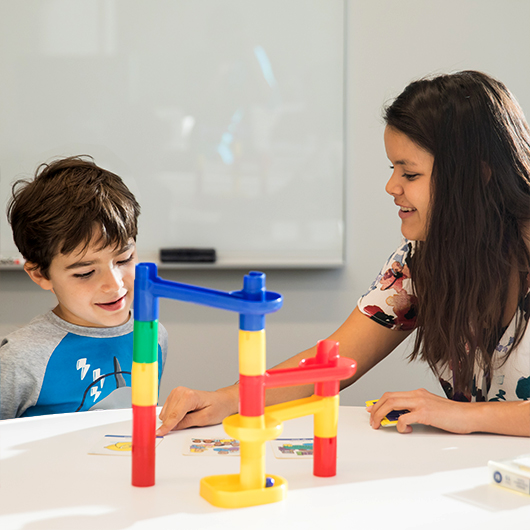 Female speech therapist works with 7-year old boy at table with colorful block toy while pointing at flash cards with 