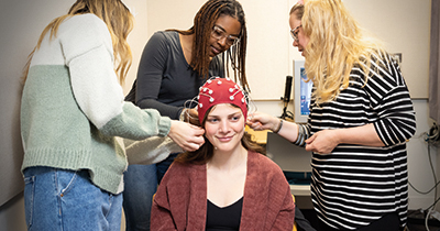 Professoer Elizabeth Norton assists students in fitting an EEG cap on student volunteer's head 