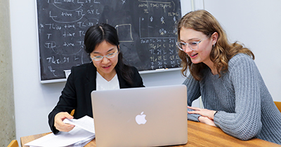 Two female students look at a laptop in front of a black chalkboard with mathematical formulas