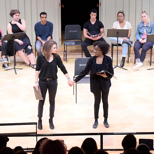 One black and one white female students clasp hands at center stage while reading lines in rehearsal with looks of consternation 