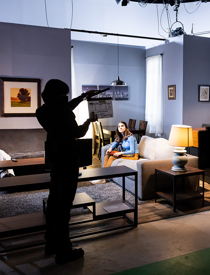 Dramatically silhouetted student holds up clapboard in front of bluish TV stage set of a living room with female actor sitting on coach