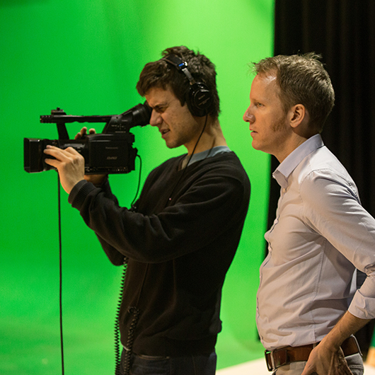 Instructor observes and directs student peering through digital video camera in front of green screen