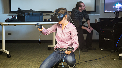 Faculty member squats to her knees while wearing virtual reality headset and controllers on each hand