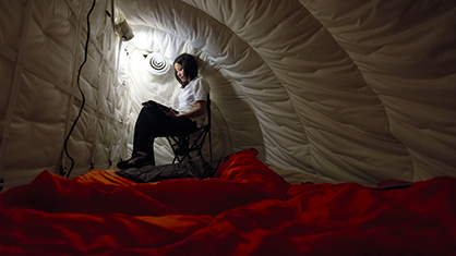 Female astronaut sits alone in padded tent room reading a book with dim light to research effects of isolation for trip to Mars