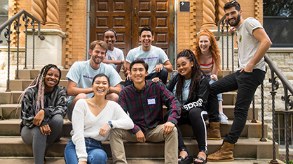 A group of nine smiling interracial students sit on the front steps of Annie Mae Swift hall