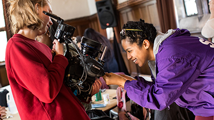 African American female in purple jacket bends down to inspect a large video camera held by a girl in red sweatshirt
