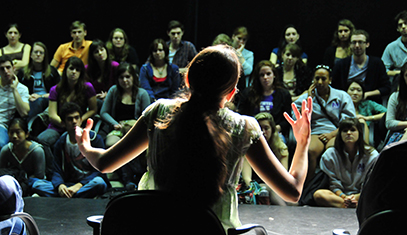 Female student sits on stage in front of crowd with her arms expressively extended wide while speaking